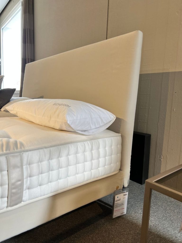 White upholstered bed with a tall headboard in a showroom, topped with a mattress and pillow, price tag hanging from the side