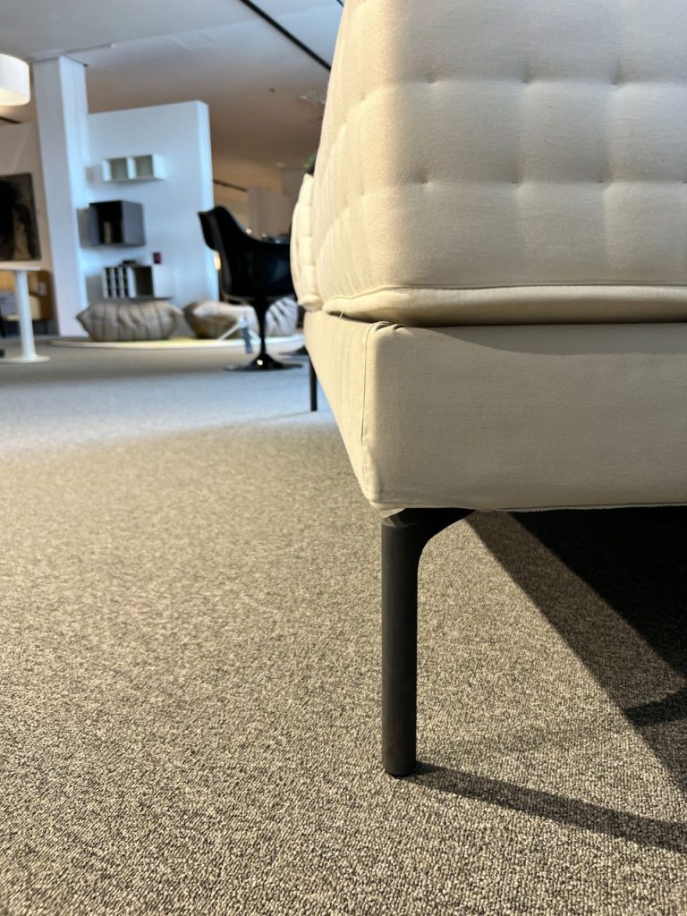 Close-up of a light beige sofa with black metal leg on a carpeted showroom floor, other furniture in background.