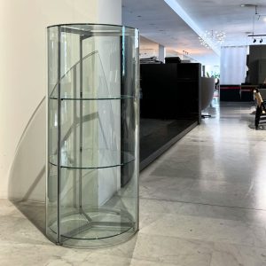 Tall cylindrical glass display case with three glass shelves in a bright showroom.
