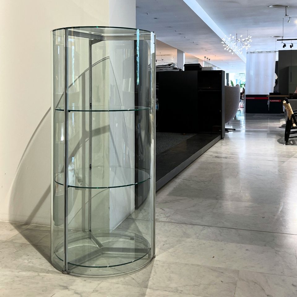 Tall cylindrical glass display case with three glass shelves in a bright showroom.