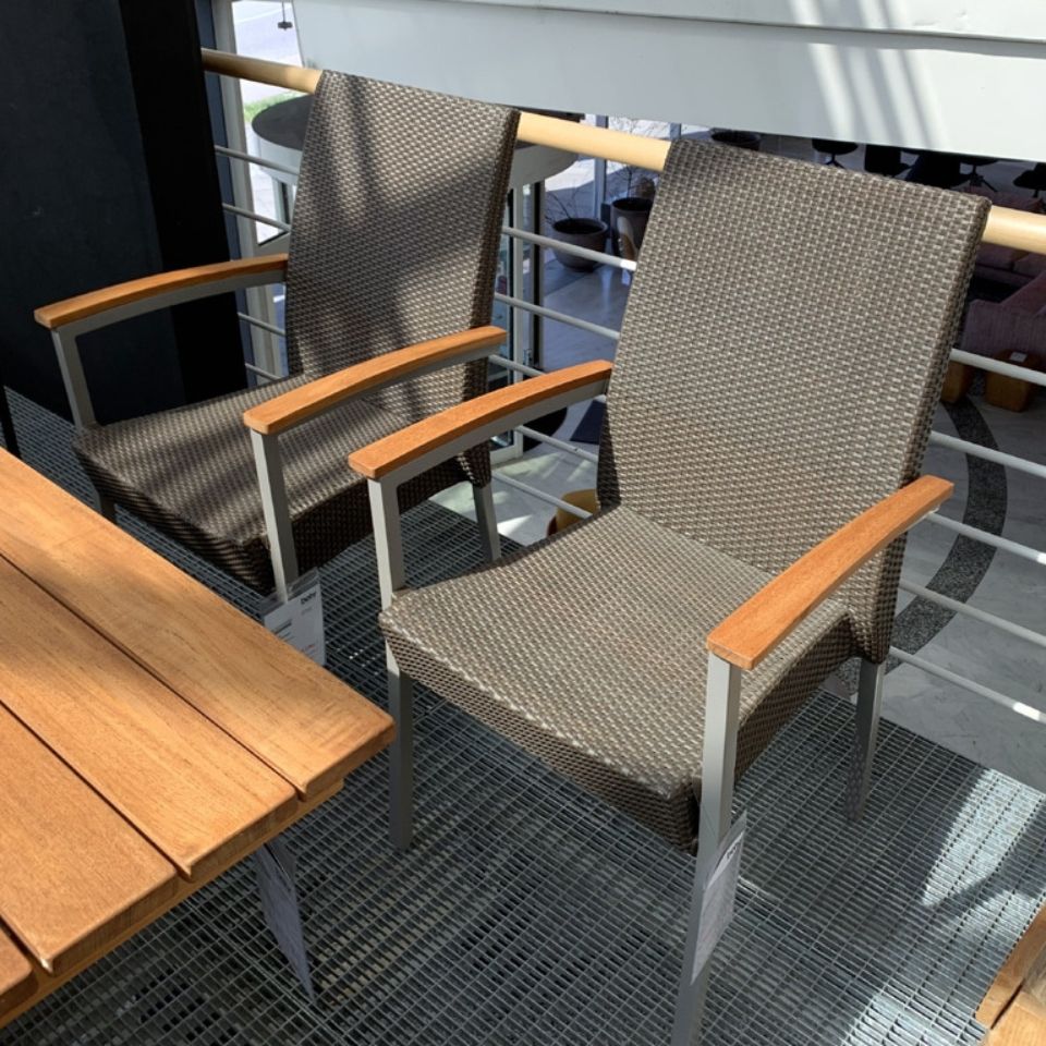 Two woven outdoor armchairs with wooden armrests on a metal grate balcony, beside a wooden table.