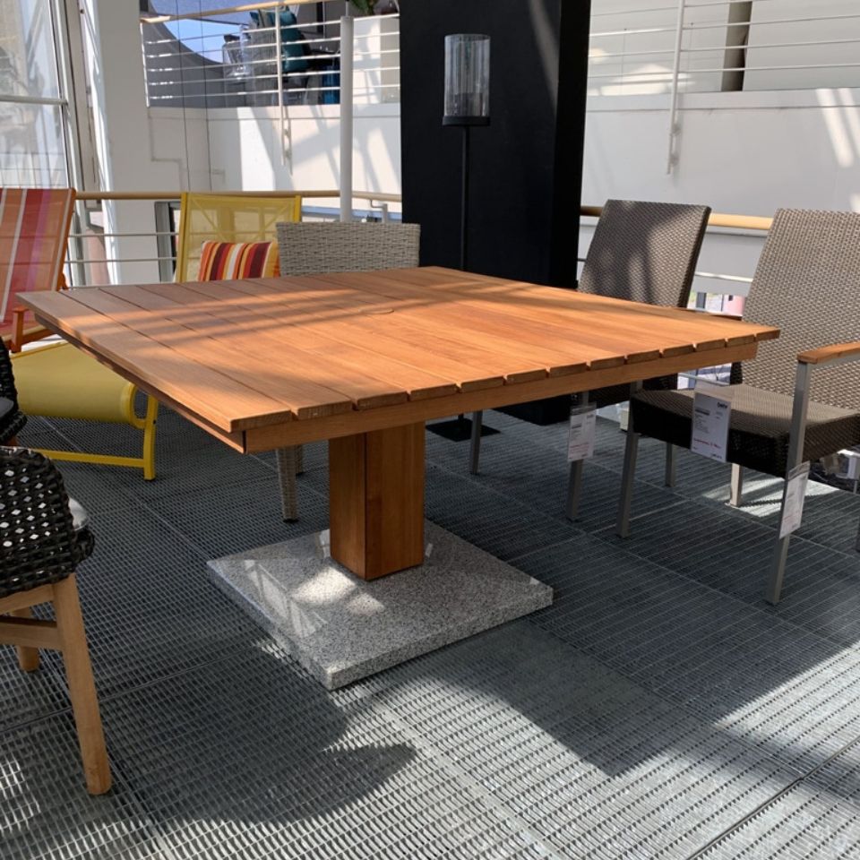 Wooden rectangular outdoor dining table with a central pedestal base, set on a granite block, surrounded by wicker and metal chairs in a bright showroom-like patio setting.