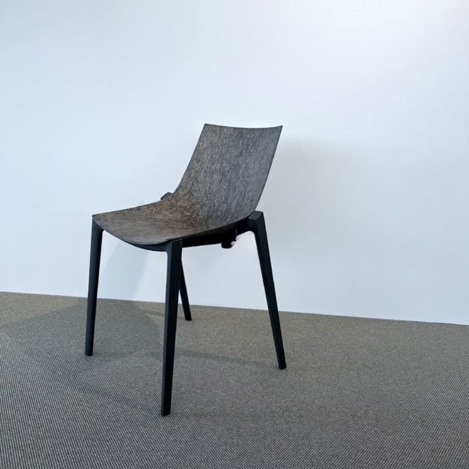 Modern dark grey curved chair with four legs on a light wall and carpeted floor.