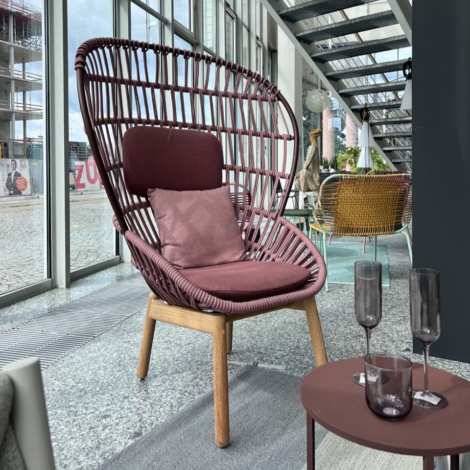 Maroon woven egg chair with a pink cushion in a modern lounge by large glass windows; a round coffee table with purple glasses sits in the foreground.