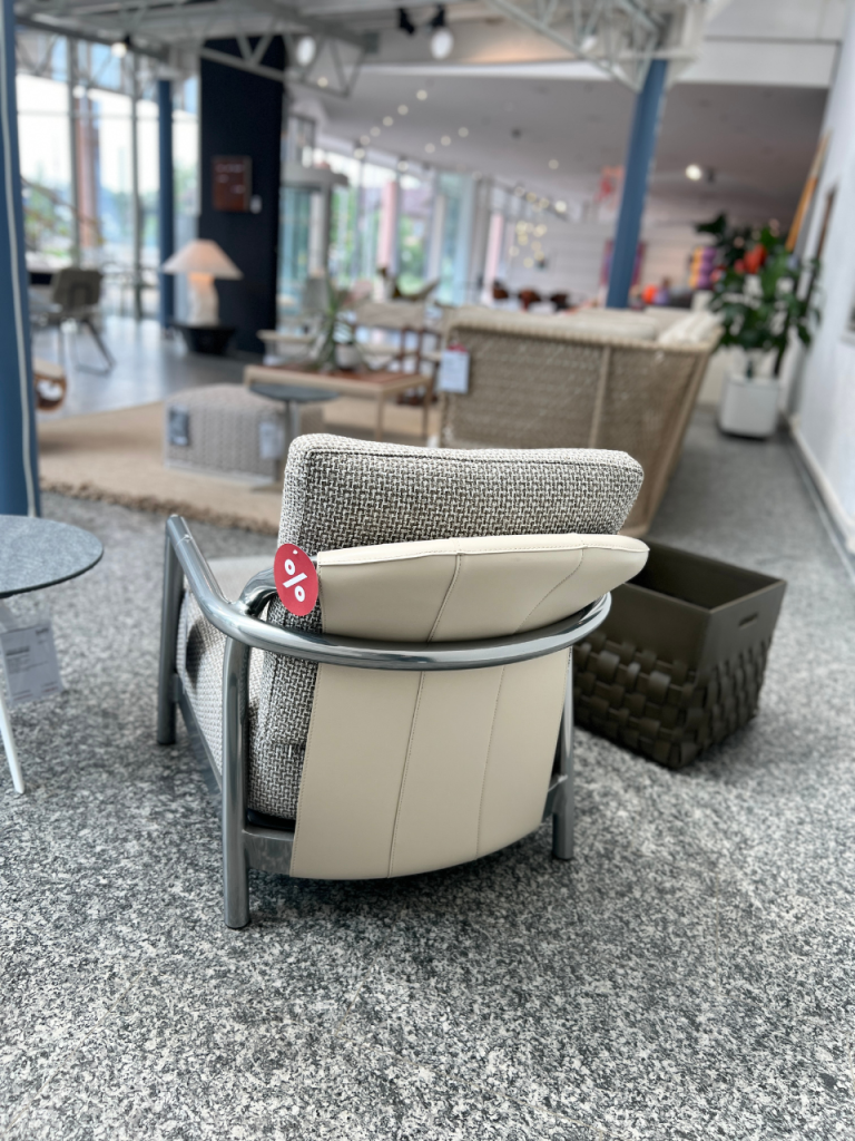 Modern showroom scene with a woven gray armchair and beige cushion in the foreground, sale tag hanging from the arm.