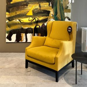 Bright yellow upholstered armchair with matching lumbar pillow in a showroom, next to a yellow floor lamp and an abstract painting on a gray wall; sale tag on chair.