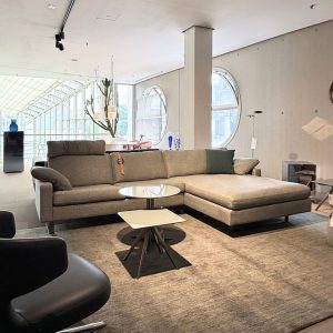 Open-plan showroom with a large gray sectional sofa, circular windows, and a glass-topped coffee table.