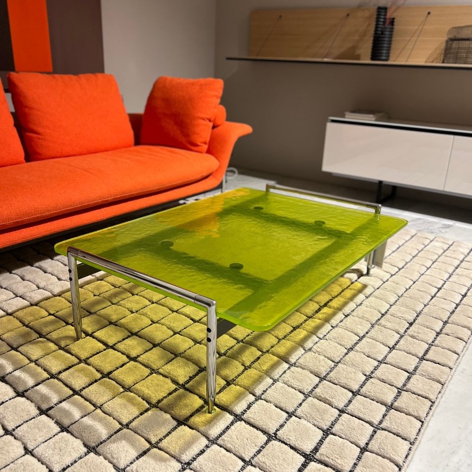 Lime-green glass coffee table with chrome legs in front of an orange sofa on a beige patterned rug.