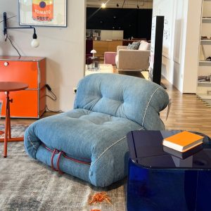 Blue tufted lounge chair with red piping in a modern showroom, beside an orange cabinet and navy glass-tinish table with an orange notebook.