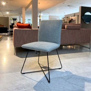 Grey upholstered chair with black metal sled legs in a showroom setting, in front of a purple sofa and modern decor.