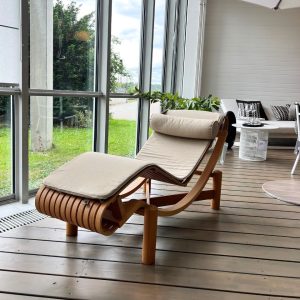 Wooden chaise lounge with beige cushions by tall glass windows in a bright indoor patio.
