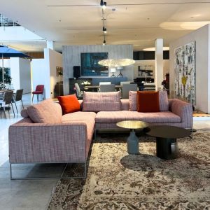 U-shaped pink fabric sofa with orange cushions in a modern showroom, centered on a patterned rug and round coffee tables.