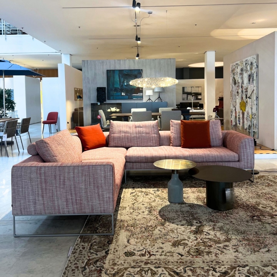 U-shaped pink fabric sofa with orange cushions in a modern showroom, centered on a patterned rug and round coffee tables.