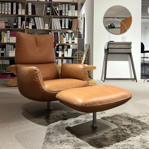 Brown leather lounge chair with matching ottoman on a gray rug in a showroom, with shelves of binders and a round-mirrored wall panel in the background.