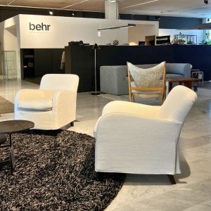 Showroom lounge with white armchairs on a dark shag rug, a black round coffee table, and a navy divider; Behr sign on the white wall in back.