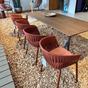 Row of rust-colored woven chairs around a long brown table in a furniture showroom with a wood-chip floor.”