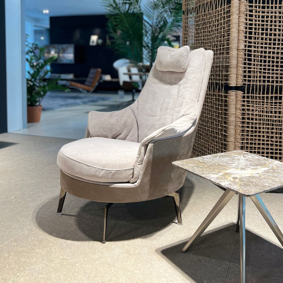 Beige tall lounge chair with cushioned headrest beside a small marble side table in a showroom setting.