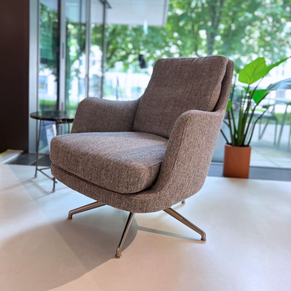 Gray upholstered swivel armchair with metal legs in a bright lounge area near a glass wall and potted plant in the background.