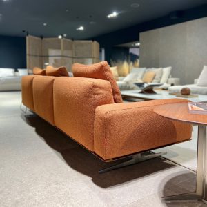 Orange textured sectional sofa with multiple cushions in a modern showroom, table nearby and other seating in the background.
