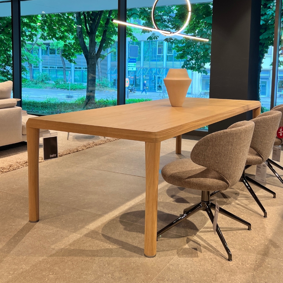 Rectangular light-wood dining table with rounded corners in a showroom, flanked by beige fabric swivel chairs; large windows and greenery outside.
