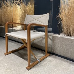 Wooden framed lounge chair with light beige cushions, placed near a glass wall with tall dried grasses in the background.