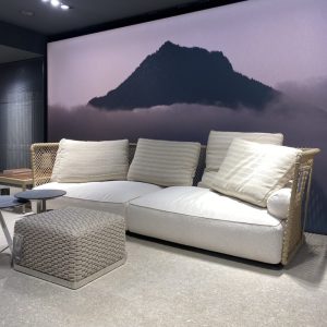 Cream woven sofa with four beige striped cushions, a matching square ottoman in front, and a large mountain mural backdrop in a showroom.