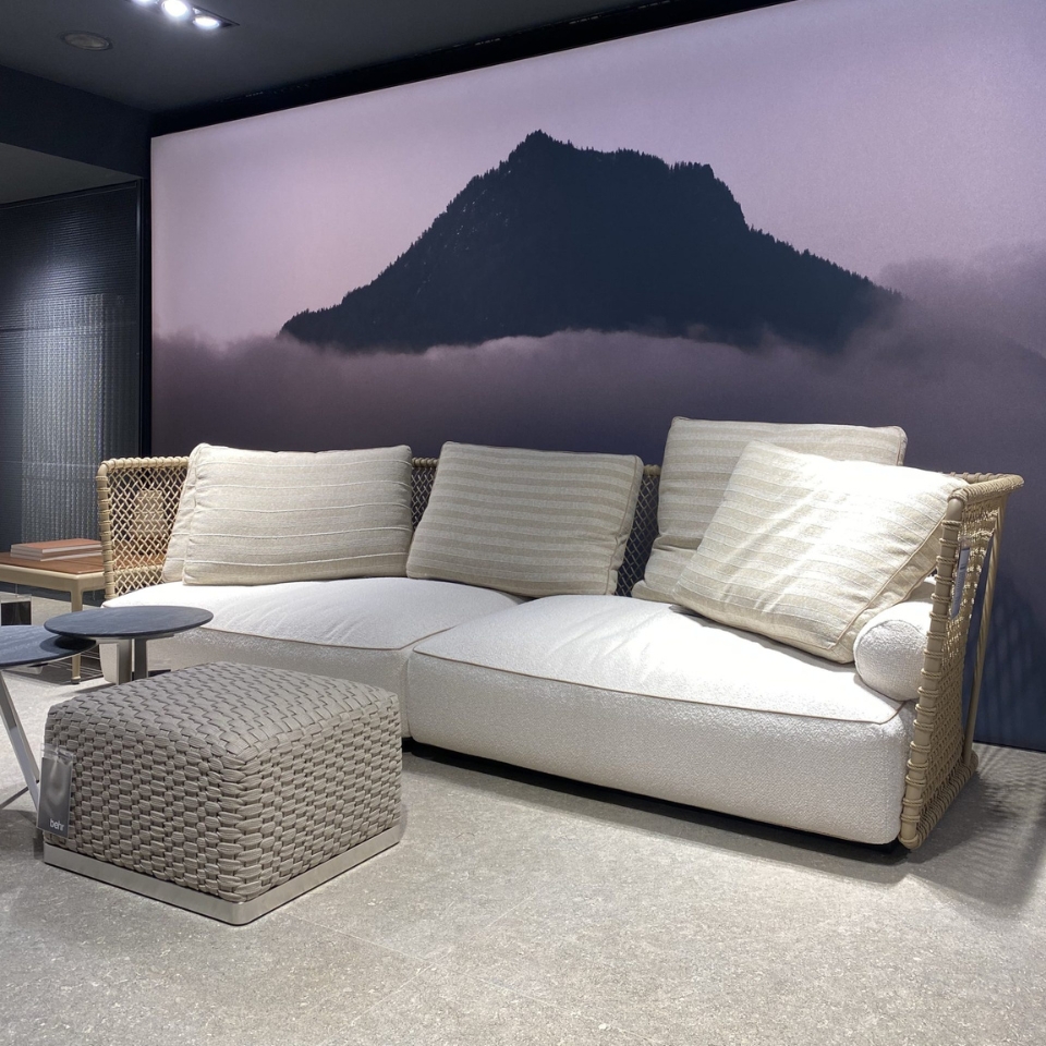 Cream woven sofa with four beige striped cushions, a matching square ottoman in front, and a large mountain mural backdrop in a showroom.