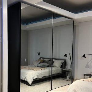 Bedroom showroom with a large black-framed mirrored wardrobe reflecting a bed with pillows and bedside tables on either side.