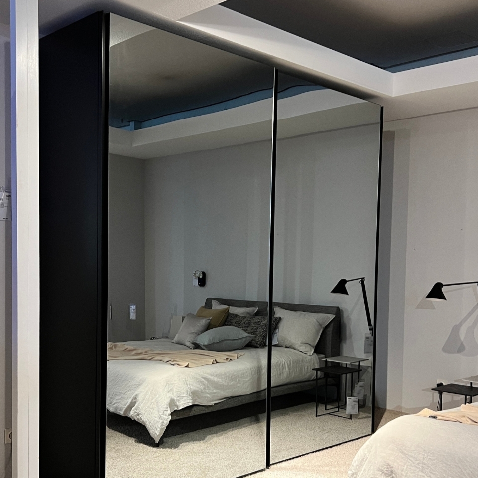 Bedroom showroom with a large black-framed mirrored wardrobe reflecting a bed with pillows and bedside tables on either side.