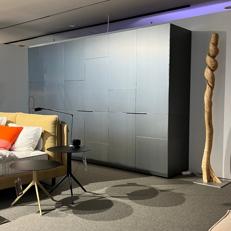 Modern showroom scene with a gray paneled storage cabinet, beige sofa with white and orange cushions, and two black round side tables on a grey carpeted floor. A twisted wooden sculpture stands at right.