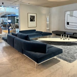 Modern showroom lounge with dark blue velvet sectional sofas around a white oval coffee table on a gray shag rug.