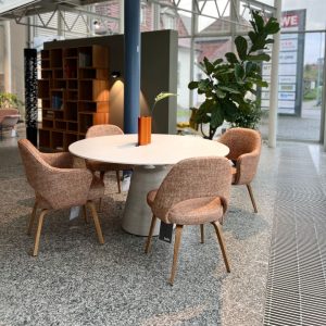 Round white table with four pink upholstered chairs in a modern showroom, orange vase with a leaf at center, large plant nearby.