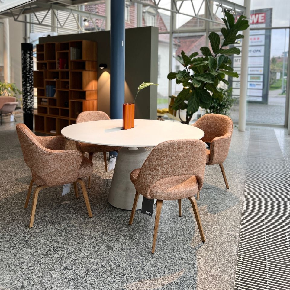 Round white table with four pink upholstered chairs in a modern showroom, orange vase with a leaf at center, large plant nearby.