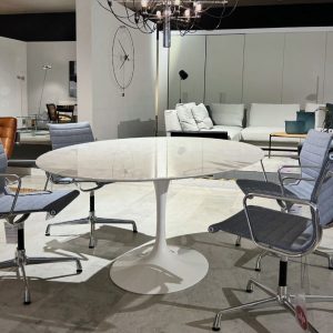 Round white marble tulip table with chrome pedestal, surrounded by four gray office chairs on a showroom floor; modern living room setup visible in the background with white sofas and a chandelier overhead.