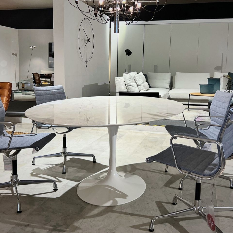 Round white marble tulip table with chrome pedestal, surrounded by four gray office chairs on a showroom floor; modern living room setup visible in the background with white sofas and a chandelier overhead.