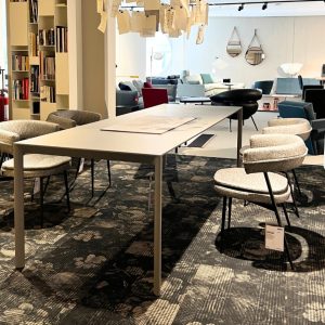 Modern showroom scene with a white table and several gray upholstered chairs around it, shelves and a lounge area in the background