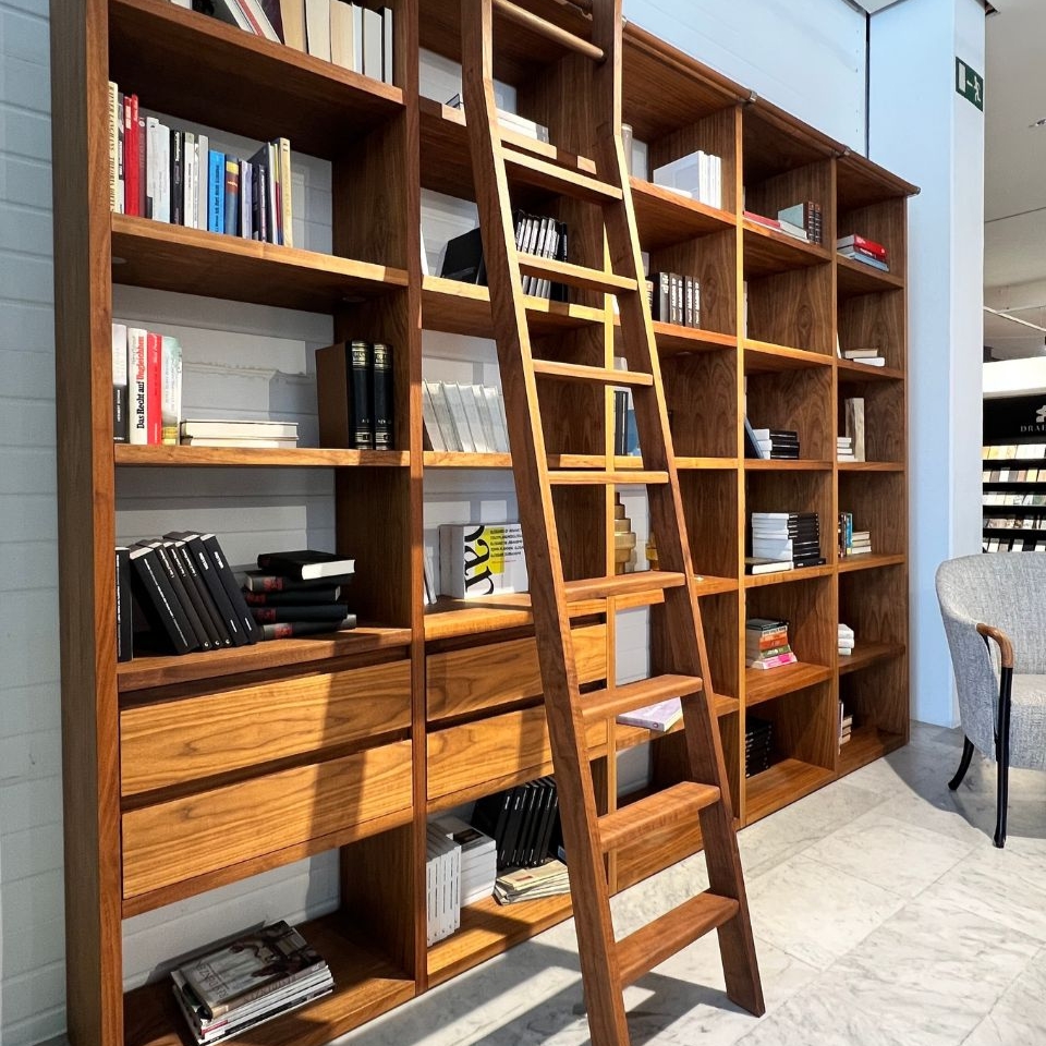 Tall wooden bookcase with multiple shelves filled with books, and a wooden ladder resting in front of it in a bright reading area.