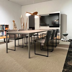 Modern conference room with a long rectangular table, gray chairs, and a large wall-mounted screen on white walls.