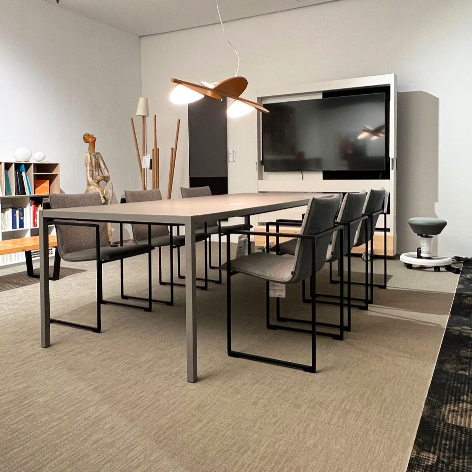 Modern conference room with a long rectangular table, gray chairs, and a large wall-mounted screen on white walls.