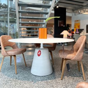 Round white table with a bright orange striped vase and a single green leaf, set in a showroom with pink upholstered chairs and a modern staircase in the background.