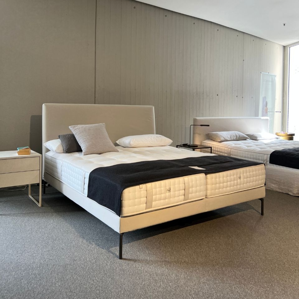 Minimalist furniture showroom with two large white upholstered beds, pillows, and a nightstand between them.