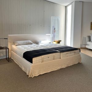 White upholstered king-size bed with tufted mattress, two pillows, beige bed skirt, and a navy throw in a showroom scene
