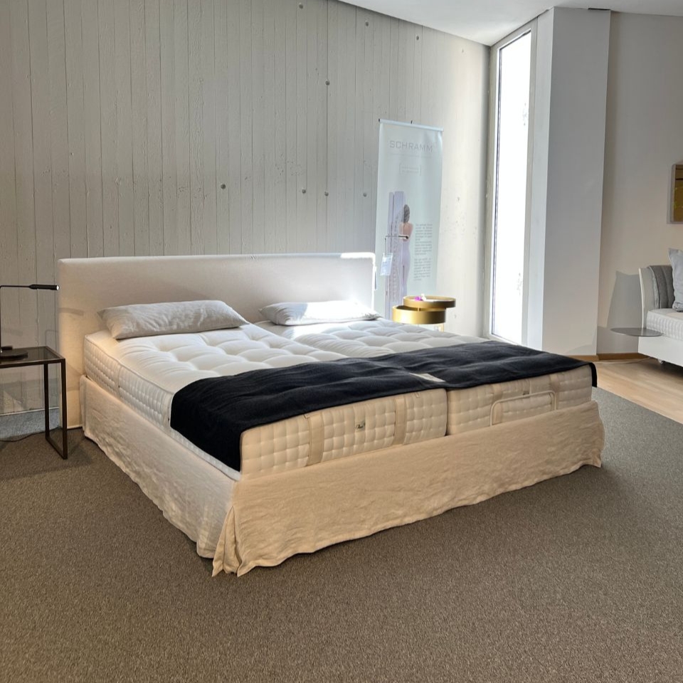 White upholstered king-size bed with tufted mattress, two pillows, beige bed skirt, and a navy throw in a showroom scene