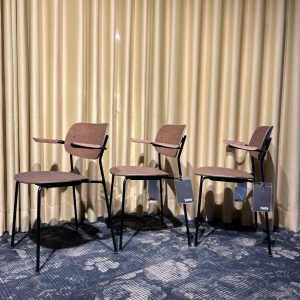 Three brown wooden chairs with attached writing tablets on metal frames, lined up in front of gold curtains.