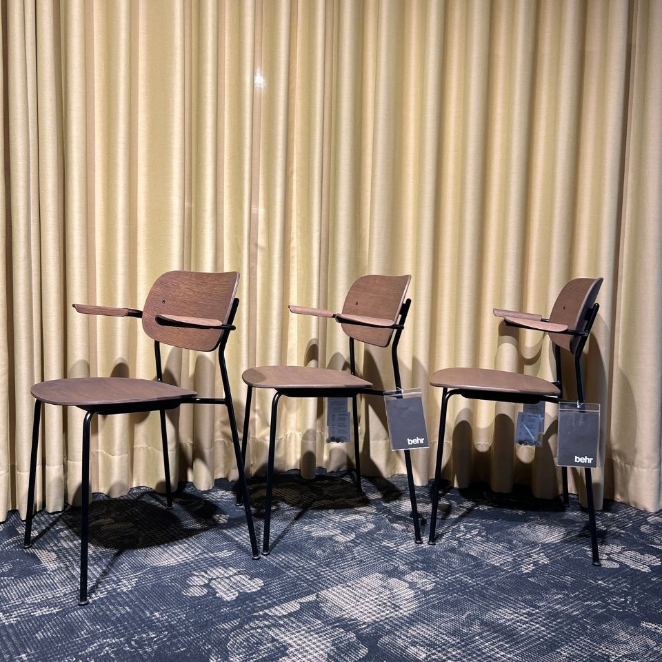 Three brown wooden chairs with attached writing tablets on metal frames, lined up in front of gold curtains.