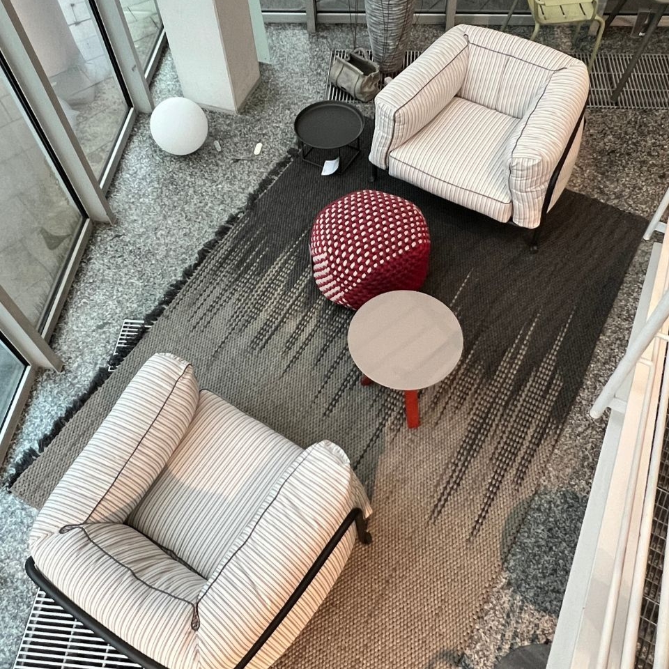 Top-down view of a lounge area with two white striped armchairs, a red pouf, a small round table, and a black side table on a striped rug.
