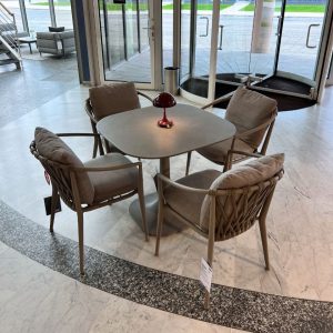 Round grey table with four cushioned wicker chairs arranged in a showroom lobby, red lamp centerpiece and glass entry doors in the background.
