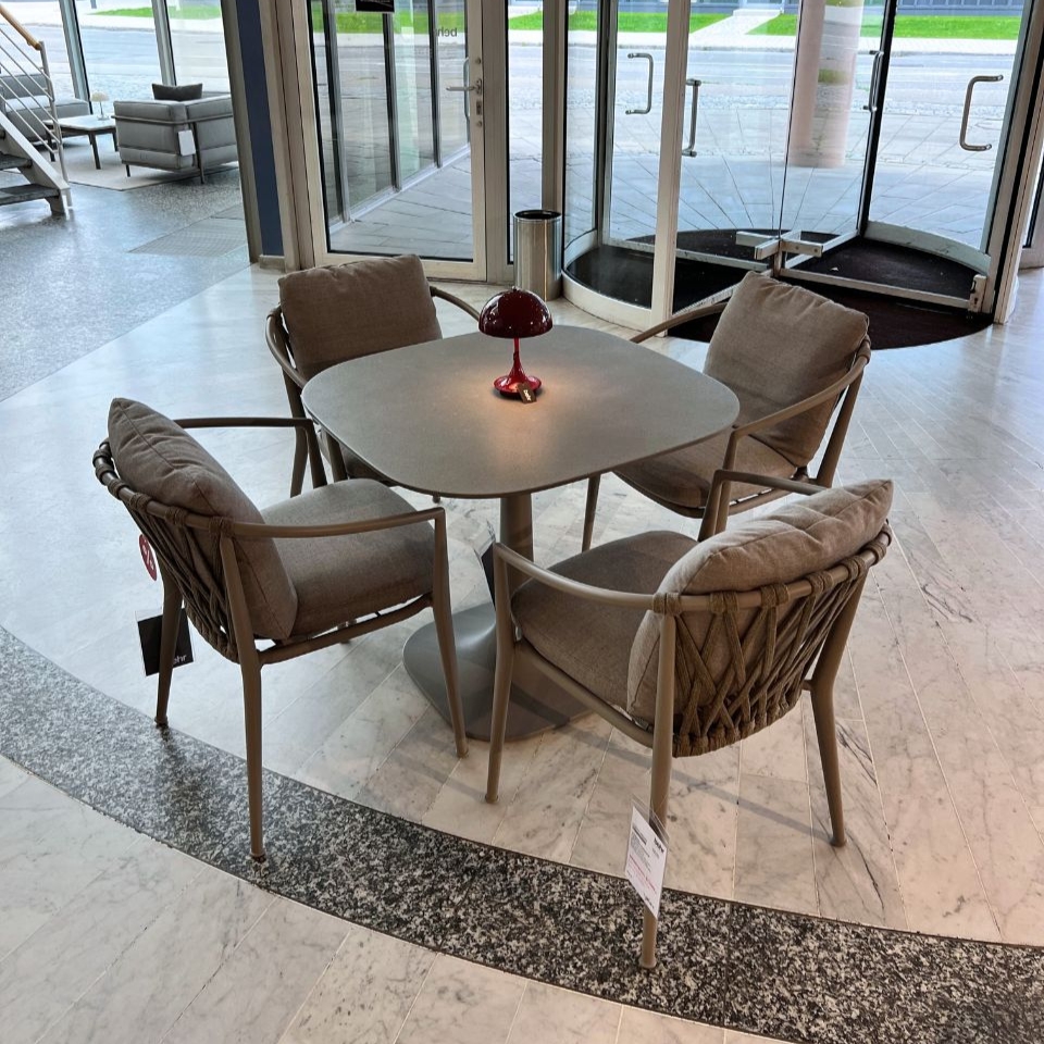 Round grey table with four cushioned wicker chairs arranged in a showroom lobby, red lamp centerpiece and glass entry doors in the background.