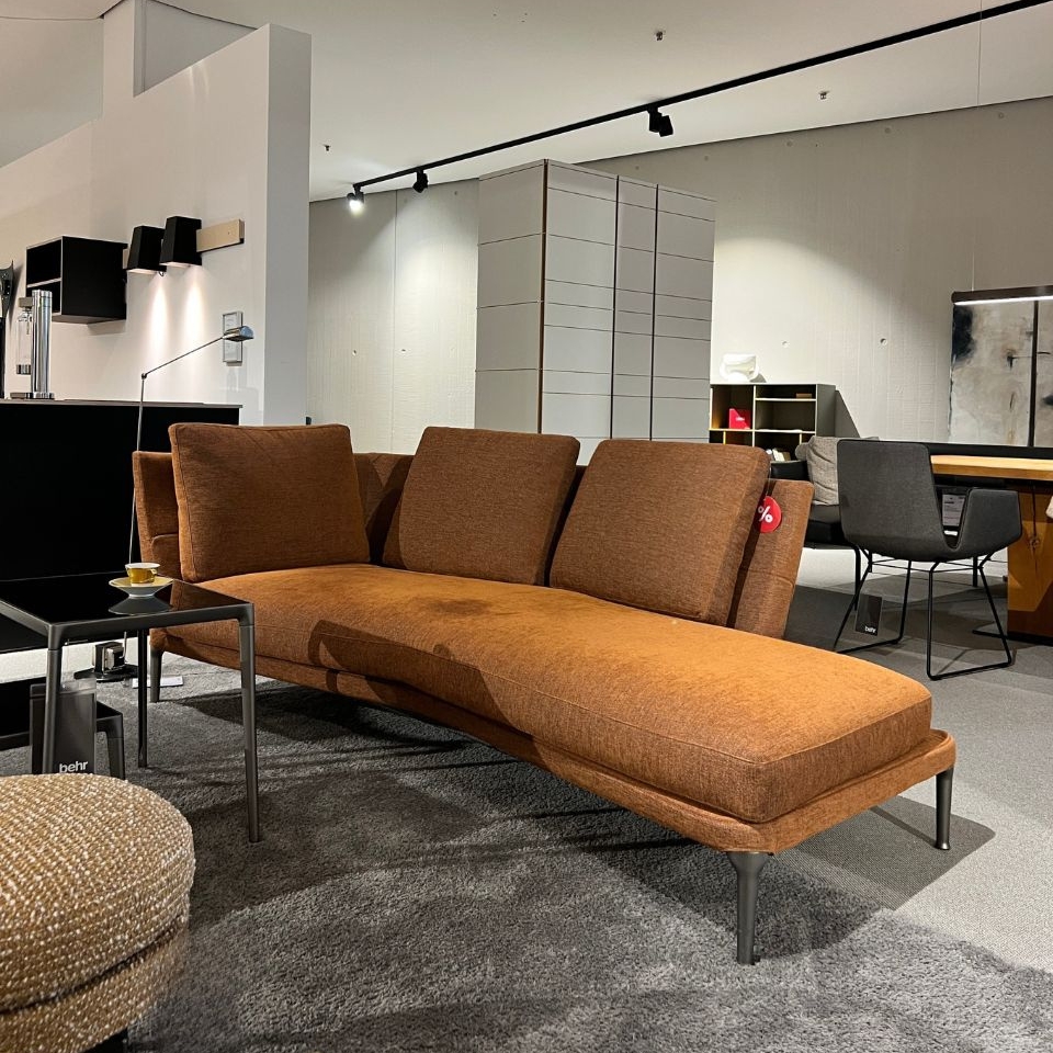 Brown fabric chaise sofa in a contemporary furniture showroom, with a small black side table and other seating in the background.