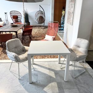 White square table with two light gray cushioned chairs on a marble floor in a bright showroom; background shows outdoor patio seating with red chairs and hanging wicker chairs under an umbrella.